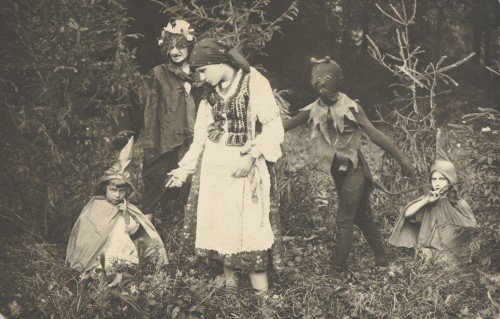 Pięcioro dzieci w kostiumach karnawałowych: dziewczynka w stroju krakowskim, chłopiec w kostiumie i masce diabła, dwoje małych dzieci w kapeluszach wróżek, dziewczynka w stroju wiedźmy ze sztucznym długim nosem, stoją i kucają w plenerze