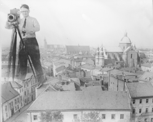 Fotografia czarno-biała, panorama Krakowa widziana z góry, dachy domów i ulice miasta. Nad jedną z ulic w formie fotomontażu umieszczona postać fotografa, stoi w rozkroku nad ulicą z aparatem fotograficznym nastatywie, w okularach, ubrany w białą koszulę, krawat w paski, czarne spodnie. Uśmiecha się i patrzy w przestrzeń 