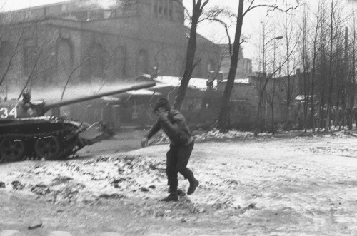 Fotografia czarno-biała. Na pierwszym planie biegnący mężczyzna w kapturze trzyma się za głowę, za nim stoi czołg. Wokół ośnieżony plac lub ulica