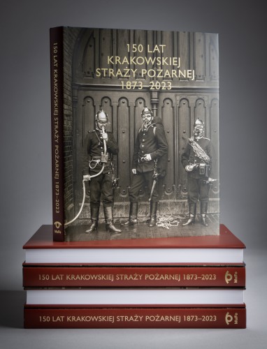 Okładka albumu „150 lat krakowskiej straży pożarnej” Książka, okładka z czarno-białym zdjęciem przedstawiającym trzech strażaków i złotym napisem, stoi na położonych na blacie czterech egzemplarzach tej samej pozycji