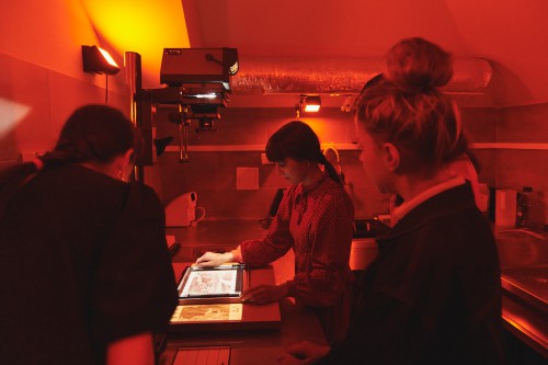 czerwone światło, trzy kobiety w ciemni fotograficznej, stoją przy działającym powiększalniku, rzucającym widok fotografii 