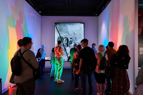 kilkanaście osób w przestrzeni wystawy, odwróceni tyłem, na wprost duża fotografia na ścianie, dookoła kolorowe światło padające na ściany