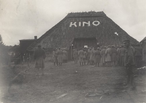 Legioniści w mundurach. Stoją w kolejce przed budynkiem krytym strzechą. Na czterospadowym dachu biały napis KINO.