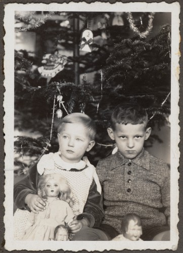 Kilkuletnie dzieci, po lewej dziewczynka z lalką, po prawej chłopiec, na tle drzewka bożonarodzeniowego. 