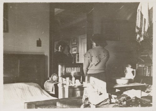 Kobieta stoi tyłem. Ubrana w strój dzianinowy z chustą w grochy. Jej twarz i fragment wnętrza widoczne w lustrze.