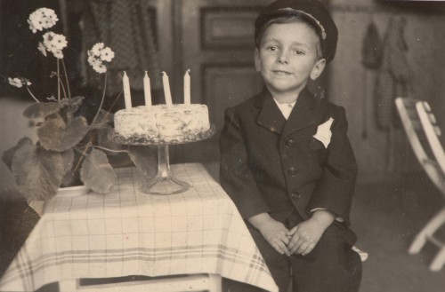 Chłopiec w ciemnym ubraniu i czapce, siedzi na krześle przy stole, na którym patera z tortem.