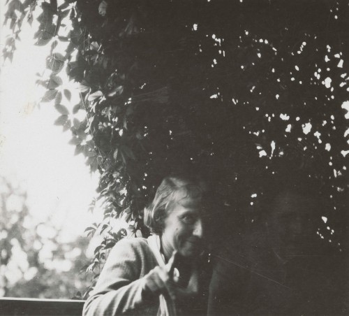 Kobieta siedząca na ganku, obok postać niewidoczna w cieniu. 