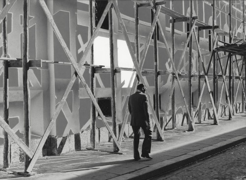 Widok miejski z przechodniem. Obok budynek osłonięty rusztowaniem.