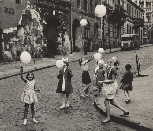 Grupa dziewczynek bawiących się balonami na brukowanej ulicy, wzdłuż której zniszczone elewacje kamienic, przy jednej z nich furgonetka.