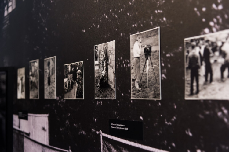Wnętrze sali wystawowej, na czarnej ścianie widoczny w jednym rzędzie ciąg siedmiu czarno-białych fotografii. Wyraźnie widoczne jedno ze zdjęć przedstawia mężczyznę stojącego przed wysokim statywem aparatu fotograficznego umieszczonym w plenerze