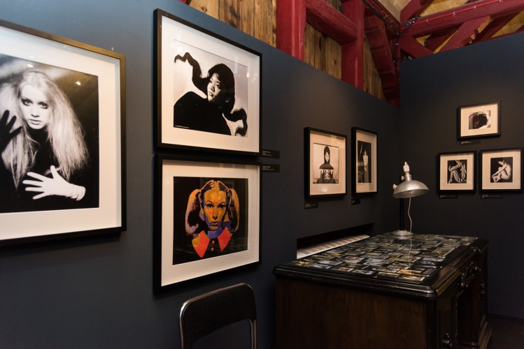 Wnętrze sali wystawowej, na pierwszym planie na ścianie w ramach fotografie przedstawiające portrety kobiet, czarno-białe i kolorowe. Po prawej stronie w rogu stoi ozdobne biurko, na blacie pod szkłem znajdują się liczne fotografie portretowe w małym formacie ułożone obok siebie